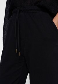 Schwarze Jogginghosen aus weichem Stoff, mit einem elastischen Bund und verstellbaren Kordeln sowie seitlichen Taschen. Dezente Markenkennzeichnung sichtbar.