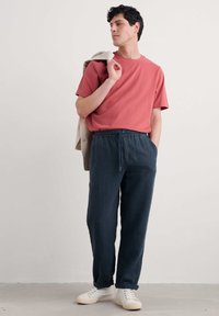 Casual outfit featuring a coral t-shirt and dark blue drawstring trousers, paired with white sneakers. Model stands with a blazer over shoulder.