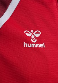 Maglia sportiva rossa con una texture a coste, con un logo bianco e il nome del marchio "hummel" sulla parte anteriore. Il design include dettagli bianchi.