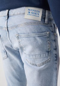Jean en denim bleu clair avec une étiquette Scotch & Soda à l'arrière, deux poches arrière et des coutures décoratives dans des couleurs pastel.
