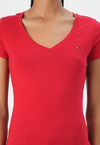 T-shirt en coton rouge avec un col en V, des manches courtes et un petit logo sur la poitrine. Texture lisse, design ajusté et sans motifs visibles.