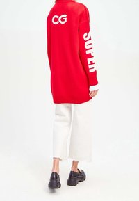 Rode trui met lange mouwen van breiwerk met "CG" en "SUPER" in het wit, gecombineerd met witte cropped broek met rafelige zoom en zwarte chunky platform schoenen.