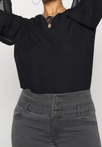 Blouse noire légère avec des manches longues transparentes et des plis. Jean en denim gris taille haute avec fermeture à bouton et poches.