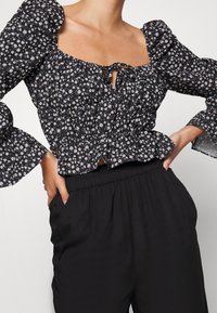 Blusa preta de flores com mangas bufantes, decote quadrado, detalhe de cordão e bainha em babados. Usada com calças largas pretas.