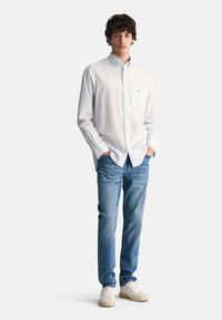 Camisa blanca de botones con cuello, jeans de color azul claro y zapatillas claras. El modelo está de pie con las manos en los bolsillos, fondo neutro.