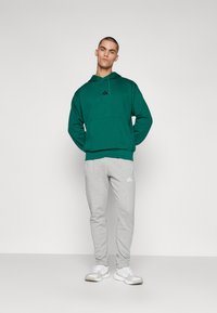 Haut de survêtement vert avec une poche avant et un logo noir, associé à un pantalon de jogging gris avec des rayures blanches et un logo, complété par des baskets blanches.