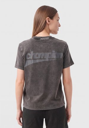 Woman facing away wearing a faded black Champion t-shirt and dark ridged shorts, standing against a plain light gray background.
