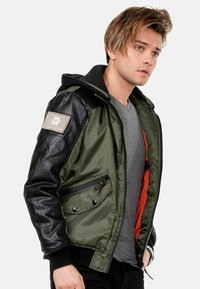 Olivgrüne Jacke mit schwarzen Lederärmeln und Kapuze, Reißverschluss und Druckknopfverschlüsse, zwei Fronttaschen und orangefarbigem Futter. Mit Aufnäher am Ärmel.