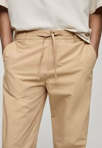 Bež hlače z elastiko v pasu in vrvico za zavezo, stranske žepke in gladko, lahko tkanino. Klasičen kroj z minimalnimi podrobnostmi.