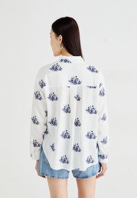 White shirt with a floral print in navy, featuring long sleeves, a button-down front, and a curved hem, paired with light denim shorts.