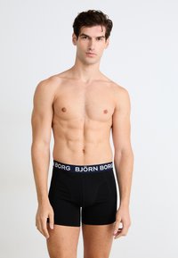 En ung mann som har på seg sorte Björn Borg boxertruser, står med et nøytralt uttrykk mot en enkel hvit bakgrunn.