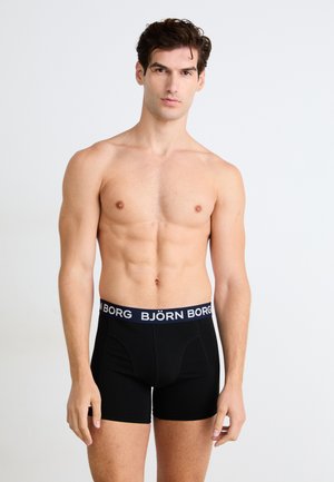 Fiatal férfi fekete Björn Borg férfi alsónadrágban, semleges arckifejezéssel áll egy sima fehér háttér előtt.