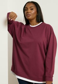 Nainen, jolla on pitkät tummat hiukset ja maroonivärinen huppari valkoisen paidan päällä, nojautuu käsivartensa valkoista seinää vasten.