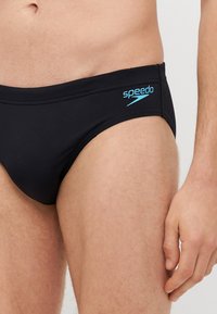 Sorte svømmebukser med blå "Speedo" logo, med elastisk talje, glat tekstur og tæt design for støtte og komfort.