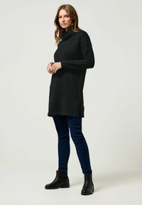 Tunic nera a maglia con collo a barchetta, aperture laterali e maniche lunghe, abbinata a jeans skinny blu e stivaletti neri.