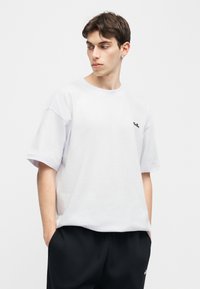 Νέος άνδρας που κοιτάζει πλαγίως, φορώντας φαρδύ λευκό μπλουζάκι με μικρό μαύρο λογότυπο και μαύρο παντελόνι, με τα χέρια στις τσέπες, απλό φόντο.