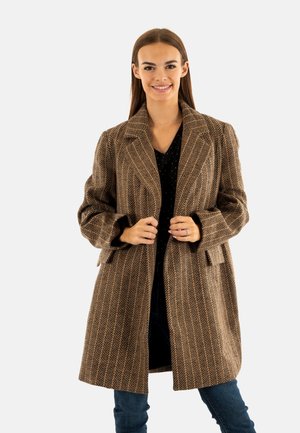 Cappotto marrone a zigzag con colletto a rever, maniche lunghe e tasche laterali, indossato sopra una maglietta nera e jeans blu.