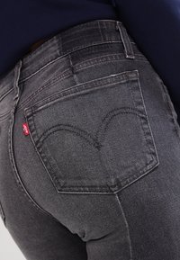Jeans de mezclilla negros con un corte de altura media, bolsillos traseros con detalles de costura y una pequeña etiqueta de logo roja, mostrando una tela texturizada.