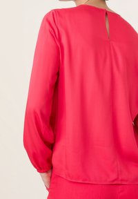 Fuchsia bluse med lange, puffede ærmer. Glat stoftekstur, med en nøglehulåbning i ryggen. Lige snitdesign med en afslappet pasform.