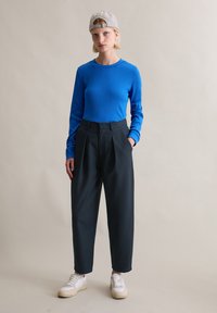 Blaues, langärmeliges, geripptes Oberteil, hoch taillierte dunkle Hose und weiße Turnschuhe. Das Model trägt eine karierte Mütze mit gebogenem Schirm.