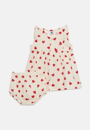 Robe bébé sans manches crème avec motif de cœurs rouges et bloomers à taille élastique assortis sur fond blanc.