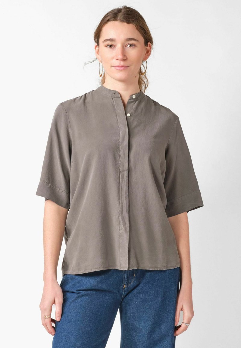 Femme portant des boucles d'oreilles créoles et une chemise grise ample boutonnée aux manches longueur coude, associée à un jean bleu, debout sur fond blanc.