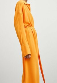 Langes orange Kleid aus einem glatten, leichten Stoff. Es hat einen Kragen, weite Ärmel und einen selbstbindenden Gürtel in der Taille.