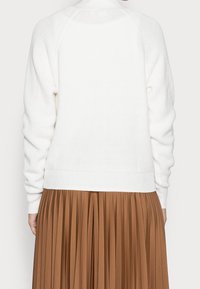 Maglione bianco lavorato a maglia con orlo e cuciture delle spalle a coste, abbinato a una gonna marrone plissettata. Visibili motivi testurizzati sul maglione.