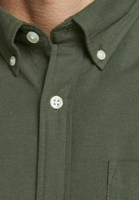 Camicia verde oliva con bottoni in tessuto testurizzato, dotata di bottoni bianchi e taschino sul petto. Colletto classico con dettagli cuciti.