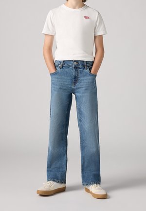 Person trägt ein weißes Levi's T-Shirt, blaue Jeans mit geradem Bein und weiße Sneakers mit Gummisohlen, Hände in den Taschen, vor grauem Hintergrund.