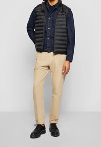 Czarna pikowana kamizelka na jeansowej koszuli, zestawiona z beżowymi spodniami i czarnymi butami. Kamizelka ma wysoki kołnierz i poziome przeszycia.