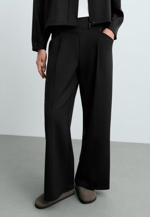 Personne portant un pantalon noir à jambes larges, une veste noire, un haut blanc et des chaussures grises sans lacets, debout sur un sol gris devant un fond clair.
