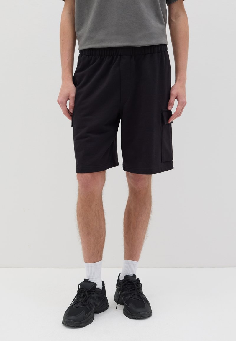 Shorts noirs avec une taille élastique, dotés d'une poche latérale ; associés à des baskets noires et des chaussettes blanches. Un T-shirt gris est visible au-dessus.