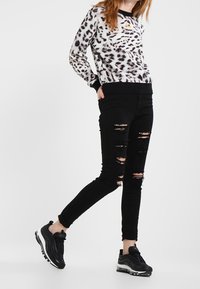 Mujer vestida con un suéter de estampado de leopardo, jeans ajustados negros rasgados y zapatillas negras, de pie con un pie ligeramente levantado.