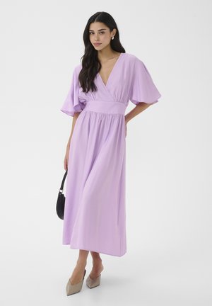 Vrouw draagt een lila wikkeljurk met brede mouwen, houdt een zwarte handtas vast, staat tegen een effen lichte achtergrond.