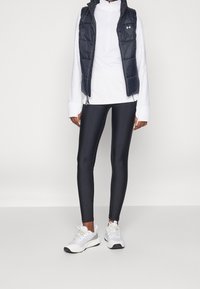 Schwarze puffy Weste über einem weißen Langarmoberteil, kombiniert mit schwarzen Leggings und weißen Turnschuhen, die ein schlankes sportliches Design aufweisen.