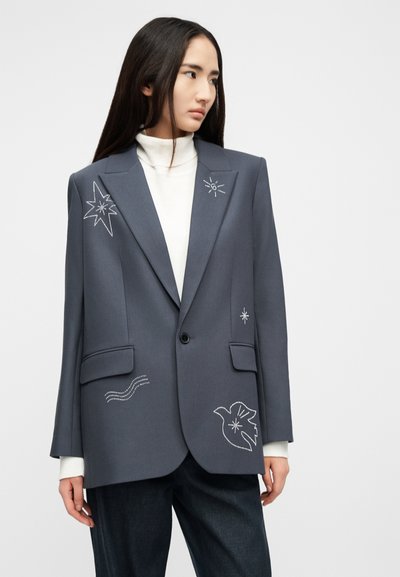 Γυναίκα που φοράει γκρι σακάκι με λευκά κεντήματα σε σχήμα αστέρα, πουλιού και κυμάτων πάνω από λευκό ζιβάγκο και σκούρο παντελόνι, κοιτάζοντας προς τα πλάγια.