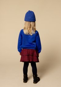 Blauwe fleece pullover, rode jurk met bloemenpatroon in lagen, zwarte maillot en zwarte laarzen, met een blauwe gebreide muts, gezien van achteren.