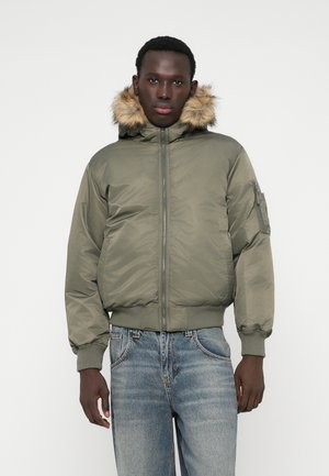 Olajzöld bomber dzseki műszőrme bélésű kapucnival, elasztikus mandzsettákkal és cipzáras elülső résszel. Oldalsó taktikai zseb található rajta.