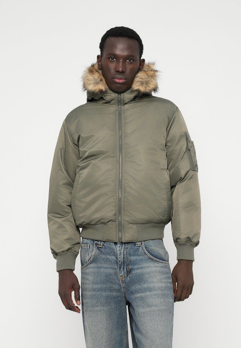 Olivroheline pommitaja jakk vale karusnahast voodriga kapuutsiga, elastsete mansettidega ja lukuga ees. Omab külgpooli utiliidikotti.