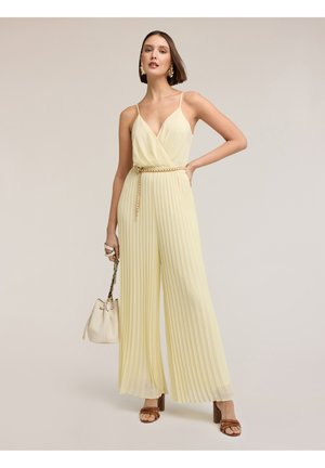 Kobieta w bezrękawniku, bladożółtym plisowanym kombinezonie z złotym łańcuchowym paskiem, trzymająca białą torebkę i nosząca brązowe sandały na obcasie.