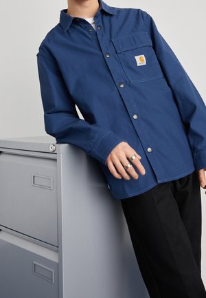 Person lehnt an einem grauen Aktenschrank, trägt ein blaues Hemd mit Knöpfen und Brusttasche sowie zwei silberne Ringe an der linken Hand.