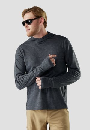 Mężczyzna w czarnych okularach przeciwsłonecznych, ciemnoszarym długim rękawie z otworami na kciuki i beżowych spodniach, patrzący w bok na jasnym tle.