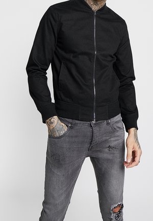 Homme portant une veste noire zippée et un jean gris déchiré, avec des tatouages visibles au cou et aux mains, debout devant un fond uni.