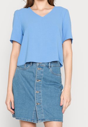 Vrouw draagt een lichtblauw blouse met korte mouwen en V-hals en een mediumblauwe spijkerrok met knopen aan de voorkant.