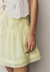 Haut blanc brodé avec une jupe jaune à volants ornée de dentelle, d'un design matelassé et d'un tissu texturé. Main posée sur la jupe.