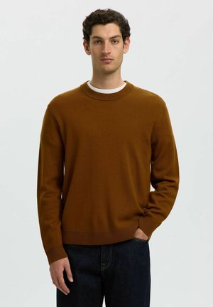 Homme aux cheveux bouclés foncés portant un pull marron à col rond sur une chemise blanche et un jean foncé, debout avec une main dans la poche.