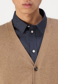 Bežový pletený cardigan s výstřihem do V a jedním tmavým knoflíkem, nošený přes námořnickou košili na knoflíky. Hladká textura, ležérní vrstvený styl.