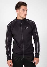 Zwarte sportjack met hoge kraag, ritssluiting en witte zijstrepen. Heeft ritszakken en geribbelde manchetten.