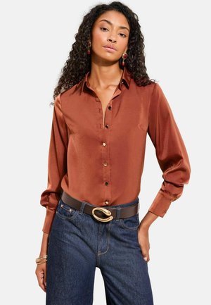 Donna con capelli ricci che indossa una blusa di raso color ruggine, jeans blu scuro, una cintura nera con fibbia dorata e orecchini rossi.